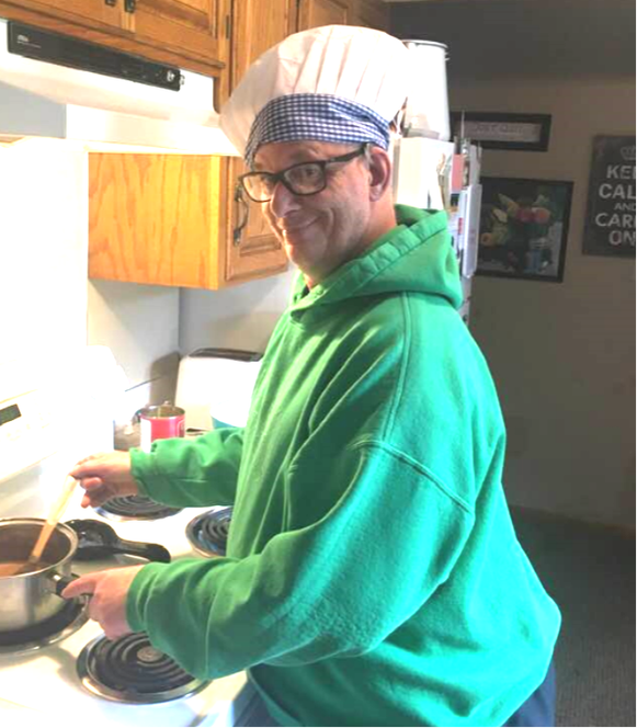 Picture of Jimmy O'Malley, a white, middle aged man wearing a green hoodie sweatshirt, black rimmed glasses and a chef's hat. Jimmy is stirring food in a pot on the stove.