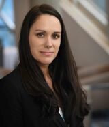 Melissa Doherty is a white woman with long black hair and brown eyes. She is wearing professional attire in head shot photo with a blurred background.