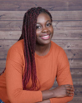 Sharlett glidden is a young black woman with long braided hair. She is wearing  an orange shirt and is posing in front of a wooden plank wall.