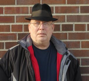 Harry is a middle-aged while male with round glasses. Harry is wearing a black fedora hat, navy shirt, and red and black winter coat. He is standing in front of a brick wall. 