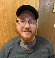 Picture of Cody Seehafer, a white male with a red beard and eye glasses wearing a ball cap. He is wearing a gray shirt and is in front of a wooden wall. 